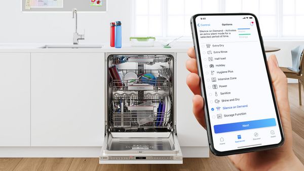 A hand holds up a smartphone showing the open Home Connect app. In the background, a dishwasher stands open revealing a full load.