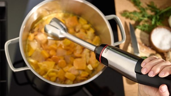 A hand holds an immersion blender over a pot of freshly cooked vegetables, ready to make smooth soup.