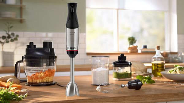 A Bosch hand blender and various accessories scattered over a wooden counter, together with a beaker of milk and other fresh ingredients.