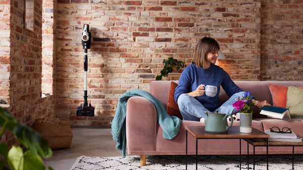 Der Unlimited 7 Akku-Staubsauger wird in der Ladestation an der Wand im Wohnzimmer aufgeladen, während eine Frau auf dem Sofa sitzt, liest und Tee trinkt.