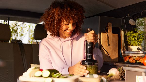 Eine dunkelhaarige Frau mit Locken und rosa Hoodie sitzt in einem Campingbus und zerkleinert Gemüse im Universal-Zerkleinerer des Bosch ErgoMaster Serie 4.