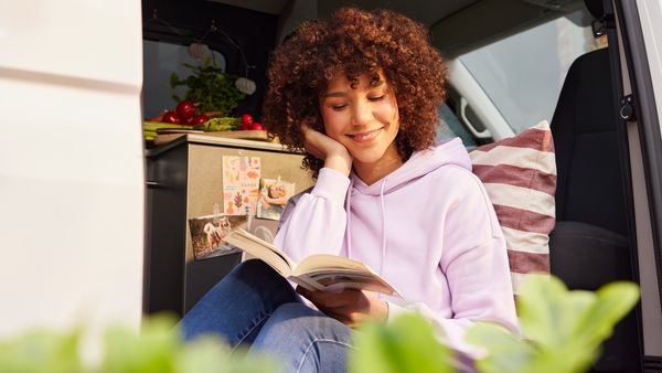 Eine dunkelhaarige Frau mit Locken und rosa Hoodie sitzt in einem Campingbus, liest ein Buch und lächelt. Sie ist an ein gestreiftes Kissen gelehnt, neben ihr ein halbhoher Schrank an den Fotos geklebt sind.