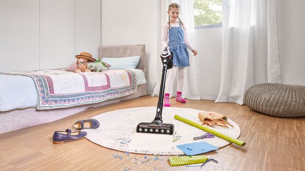 A young girl using a vacuum cleaner to clean her room, with toys scattered on the floor.