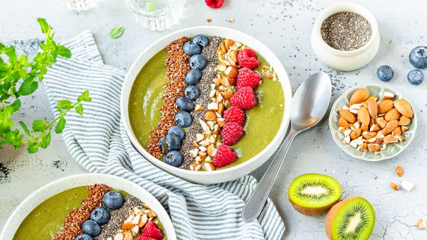 Sklede zelenih smoothiejev, obložene z borovnicami, malinami, oreščki in semeni, z rezinami kivija in kozarcem semen chia na teksturirani sivi površini.