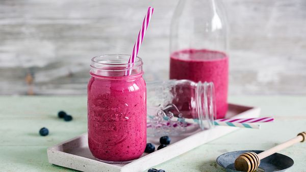 Živahen roza jagodni smoothie v kozarcu s črtasto roza slamico, obdan z borovnicami in medeno zajemalko na rustikalnem lesenem ozadju.