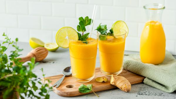 Dva kozarca mangovega smoothieja z limeto in meto, vrč smoothieja in svež ingver na pultu.