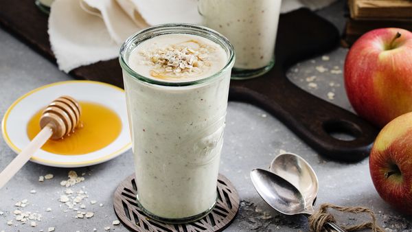 Kremni jabolčni ovseni smoothie z ovsenimi kosmiči, postrežen v kozarcu z medeno posodico in svežimi jabolki na sivi teksturirani površini.