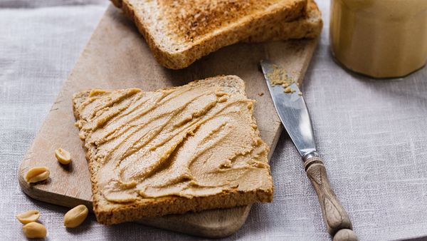 Кусок тоста со сливочно-арахисовым маслом, помещенный на деревянную доску с ножом и арахисом рядом с ним.