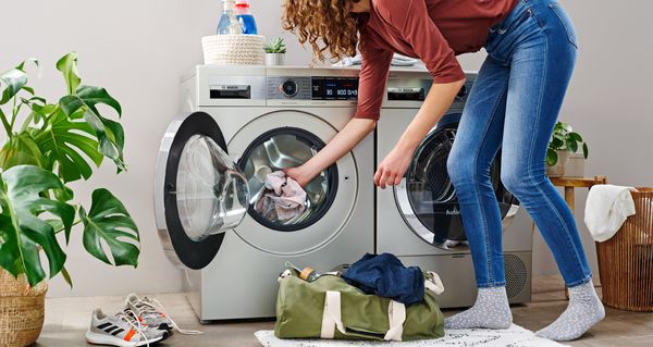 Eine Frau in Blue-Jeans und rotem Oberteil gibt Sportsachen aus ihrer Sporttasche in eine Waschmaschine von Bosch.