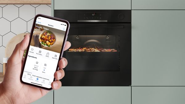 Op de voorgrond houdt een hand een smartphone omhoog met daarop een recept voor groentechips in de Bosch Home Connect-app, terwijl de ingrediënten in de oven op de achtergrond garen.