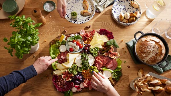 Zu sehen ist ein Charcuterie Board von oben, an dem sich gerade mehrere Personen mit verschiedenen Leckereien bedienen.