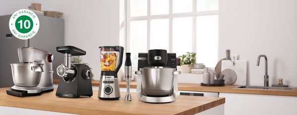 Robot de bucătărie OptiMUM Serie 8, robot de bucătărie Serie 6, blender  de masă VitaPower Serie 4 și blender de mână ErgoMaster Serie 6 stand pe un blat, lângă simbolul de garanție 10 ani.