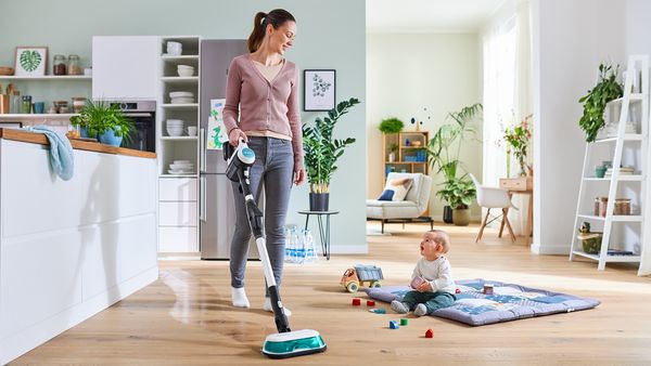 En ung kvinne bruker ProHygienic Aqua mop og støvsuger til å rengjøre tregulv i et lys stueområde. En baby leker på et teppe midt i rommet.