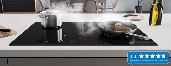 Table de cuisson Bosch avec module de ventilation intégré. Une poêle avec un steak et une casserole au premier plan, une salle à manger avec une table en arrière-plan.