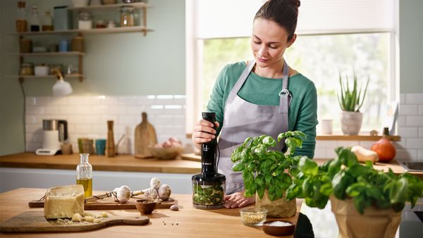 Eine Frau steht in einer Küche und mixt Kräuter mit einem Ergomaster Stabmixer von Bosch, um frisches Pesto zuzubereiten.