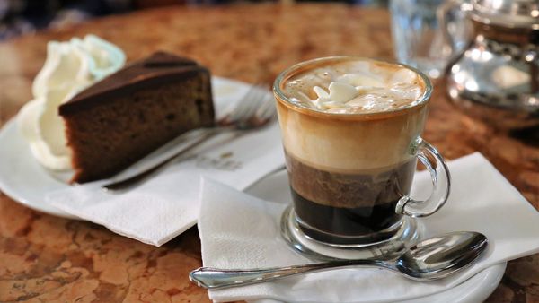 A cup of Wiener Mélange coffee beside a slice of cake on a wooden table. 