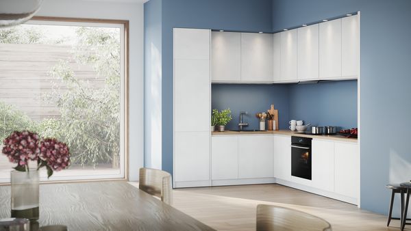 Una cocina moderna con armarios blancos, una pared azul y una encimera de madera; cerca hay una mesa de comedor con flores.