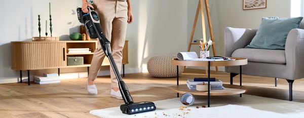 Eine Person in beiger Hose reinigt mit einem Bosch Unlimited 10 Akku-Staubsauger einen weißen Teppich, auf dem eine umgefallene Schüssel vom Tisch Schmutz verursacht hat.