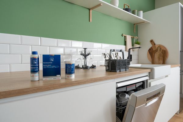 I ett kök med gröna väggar står olika rengöringsprodukter och tillbehör för diskmaskiner från Bosch på en köksbänk. Bredvid en öppen diskmaskin syns maskinrengöring, en bestickkorg och en glashållare.