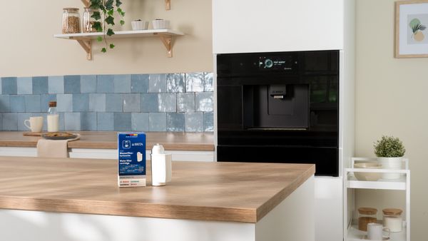 Une cuisine blanche avec un four et une machine à café Bosch encastrés en arrière-plan, et un filtre à eau Brita posé sur le plan de travail. Sur l’image, un macaron bleu avec le texte : Achetez votre pack maintenant. 