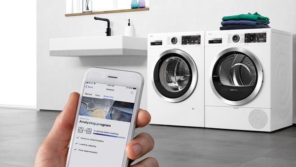 A hand holds a smartphone, showing the Bosch Home Connect app, which operates the washing machine and dryer in the background.