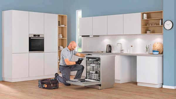 Technician repairs a Bosch dishwasher in a blue kitchen.