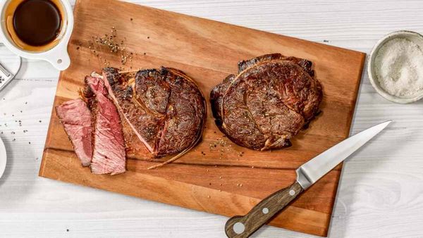 Tagliere di legno con due bistecche cotte e una fetta di carne affettata, accompagnate da una forchetta e un coltello. 