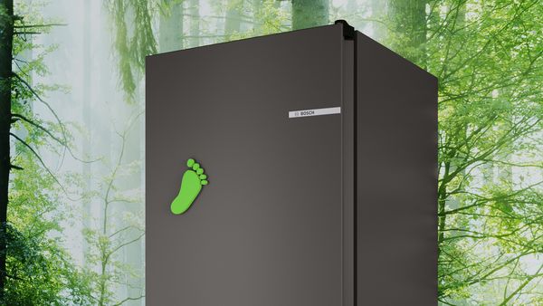 Ein dunkelgrauer Kühlschrank in Nahaufnahme mit einem grünen Fussabdruck-Magneten. Im Hintergrund ist eine Tapete mit einem grünen Wald zu sehen.