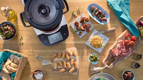 Die Bosch Cookit Küchenmaschine, die kocht, steht auf einem Tisch voller Tapas wie Oliven, Brot und Schinken.