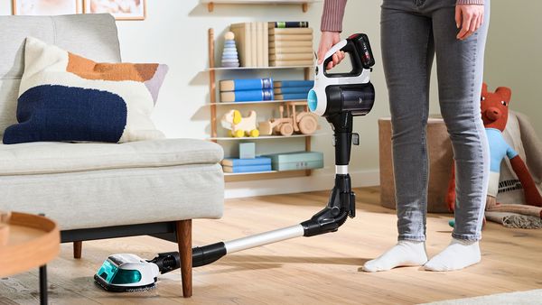 Na obrázku je osoba, ktorá používa bezdrôtový vysávač na čistenie podlahy pod pohovkou. V obývacej izbe sú hračky a knižky, čo naznačuje prítomnosť detí. Izba vyžaruje moderný a útulný pocit.     