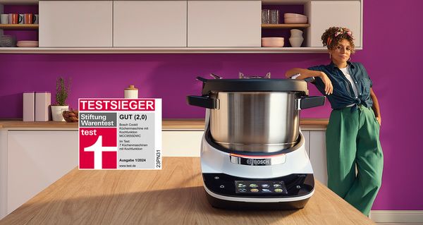 In einer modernen Küche steht die Bosch Cookit Küchenmaschine, die kocht, auf der Arbeitsplatte. Rechts lehnt eine Frau am Tresen, vor einer lila Wand im Hintergrund. Ein Testsiegel von Stiftung Warentest (Ausgabe 1/2024) bestätigt den ersten Platz des Bosch Cookit unter sieben Küchenmaschinen mit der Note "GUT (2,0)".