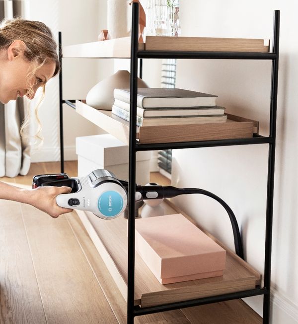 A woman vacuums a shelf, removing dust and debris with a handheld vacuum cleaner. 