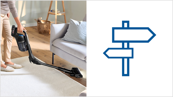 Kabelloser Stielsauger an Teppichkante im Wohnzimmer; daneben Wegweiser-Symbol.