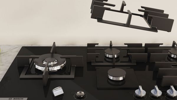 Vue de face de la table de cuisson gaz. Un bouton est retiré et placé sur la plaque et un support de casseroles est suspendu.