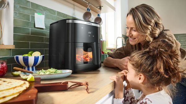 Una madre e una figlia osservano entusiaste il cibo in cottura in una friggitrice ad aria Bosch Serie 6 dalla finestra di controllo. La cucina è moderna e luminosa.