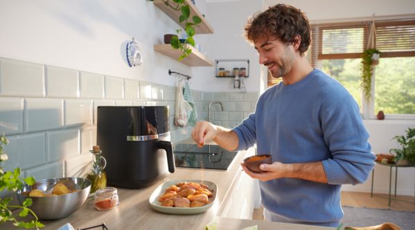 Um homem numa cozinha luminosa, segura uma pequena tigela de temperos e polvilha a mistura de especiarias sobre a carne crua no balcão. A Air Fryer Serie 4 da Bosch está no balcão ao fundo.