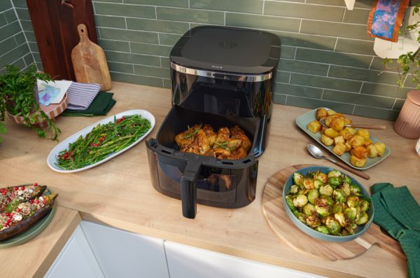 Friggitrice ad aria aperta su un piano cucina con pollo nel cestello, circondata da contorni di verdure e patate arrosto