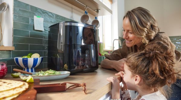 Uma mãe e a sua filha, ambas a observar ansiosamente através da janela de visualização de uma Air Fryer Bosch Serie 6, enquanto os alimentos cozinham no interior. A cozinha é luminosa e moderna.