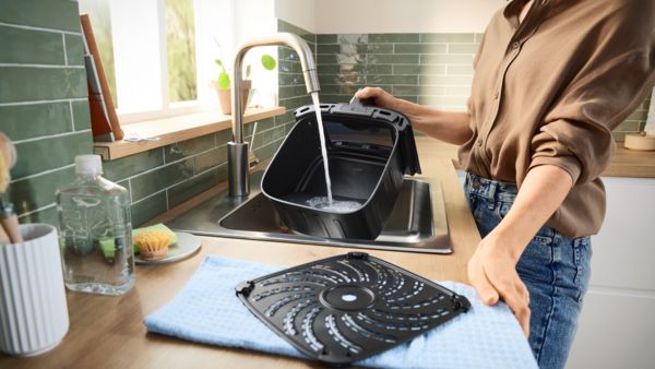 Uma mulher lava o cesto da fritadeira Bosch Air Fryer sob a torneira do lava-loiça enquanto a grelha amovível seca sobre um pano de cozinha na bancada da cozinha.