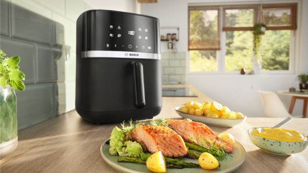 Um prato com filetes de salmão tenros, espargos estaladiços e batatas assadas douradas está em frente a uma Air Fryer da Bosch numa bancada de cozinha, mostrando as capacidades de eficiência energética e de cozedura rápida do eletrodoméstico.