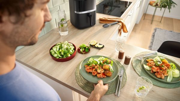 Um jovem coloca um prato de comida acabada de cozinhar no balcão da cozinha. Ao lado, um segundo prato e uma tigela de salada de pepino.