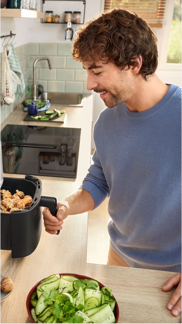 Um jovem numa cozinha, puxando parcialmente o cesto da sua fritadeira de ar quente para verificar se os alimentos estão cozinhados.