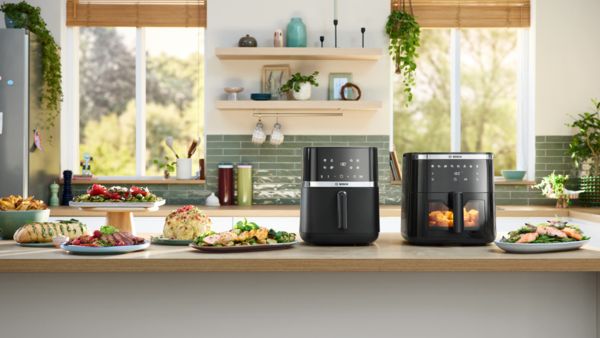 Duas Air Fryers da Bosch, Serie 4 e Serie 6, colocadas lado a lado numa ilha de cozinha, com pratos vegetarianos e de carne dispostos nas proximidades.