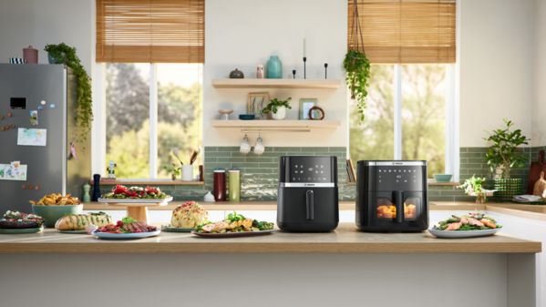 Duas Air Fryers Bosch posicionadas lado a lado num balcão de cozinha luminoso. Vários pratos e tigelas de comida, incluindo pão de ervas, frango, assado e batatas fritas, são colocados ao lado dos aparelhos, mostrando pratos confecionados com as Air Fryers.