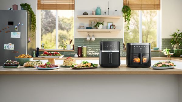 Dois modelos de Air Fryer Bosch Serie 4 e Serie 6, colocados lado a lado numa ilha de cozinha, com pratos vegetarianos e de carne dispostos à sua volta.