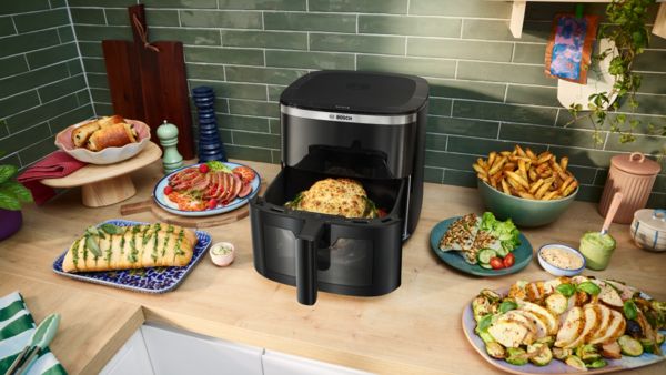 A gaveta aberta de uma Air Fryer Serie 6 da Bosch mostra frango acabado de cozinhar. Ao lado, legumes cozinhados como batatas, couves-de-bruxelas, espargos e beringelas repousam no balcão.