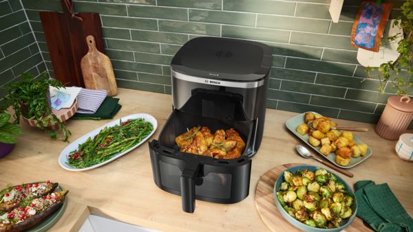 A gaveta aberta de uma Air Fryer Serie 6 da Bosch mostra frango acabado de cozinhar. Ao lado, legumes cozinhados como batatas, couves-de-bruxelas, espargos e beringelas repousam no balcão.