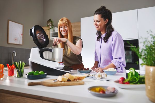 Due donne sono in cucina accanto al Cookit, una aggiunge un ingrediente.