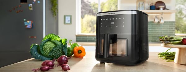 Une friteuse &agrave; air Bosch s&eacute;rie 6 est pos&eacute;e sur un comptoir de cuisine entour&eacute;e de l&eacute;gumes frais dans une cuisine lumineuse et moderne, pr&ecirc;te &agrave; cuisiner.