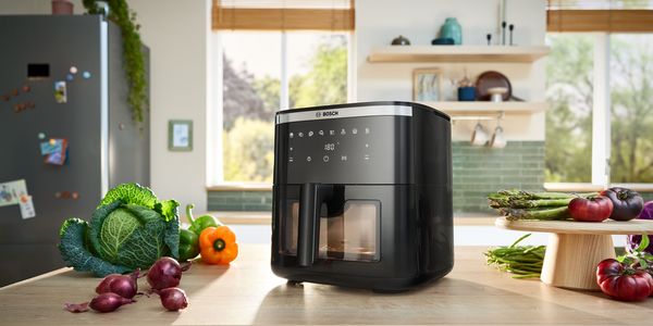Uma Air Fryer Serie 6 está no balcão da cozinha rodeada de vegetais frescos numa cozinha moderna e luminosa, pronta a cozinhar.